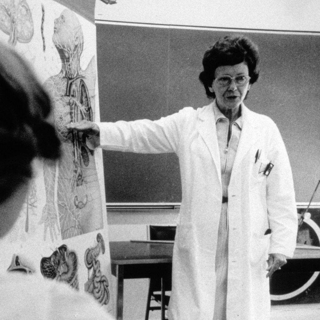 Vera Peters, in a lab coat, stands in front of a classroom and points to a flip chart featuring anatomical illustrations.