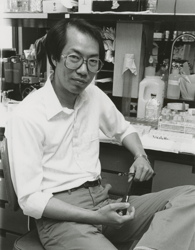 Tak Mak leans casually on a lab bench that is covered with scientific equipment.