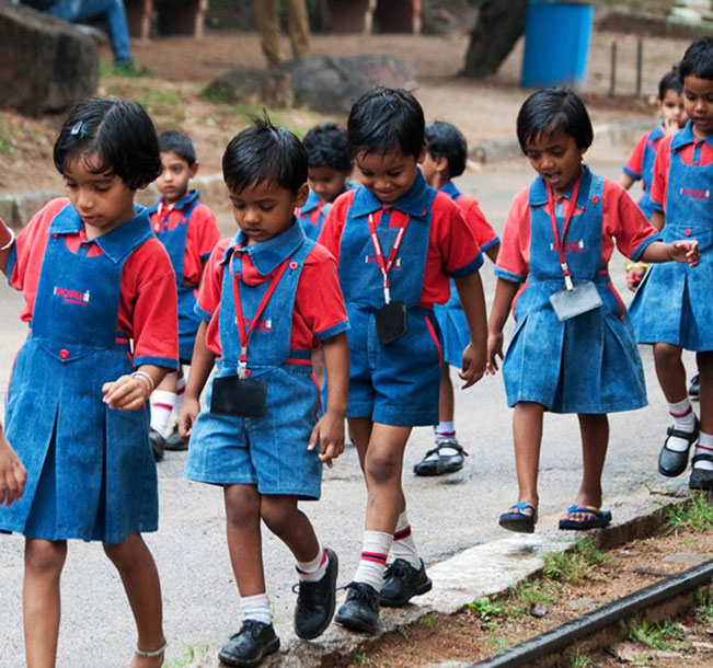 Group of young school children in India. They are dressed in red and blue uniforms and are walking in a line on a paved road.
