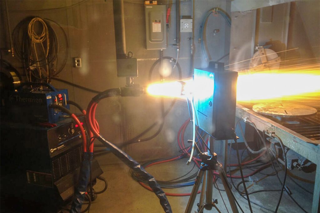 In a lab, a jet of something bright and hot shoots out of a hose and through a metal box.