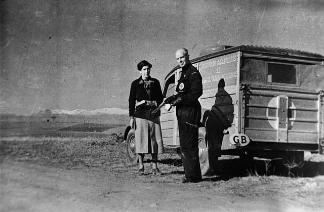 A woman and a uniformed man who is likely Norman Bethune stand in a desert beside a truck painted with the Red Cross symbol.