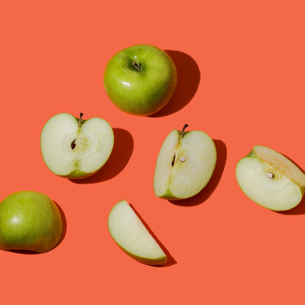 A whole granny smith apple and some apple slices sit on a salmon-coloured background.