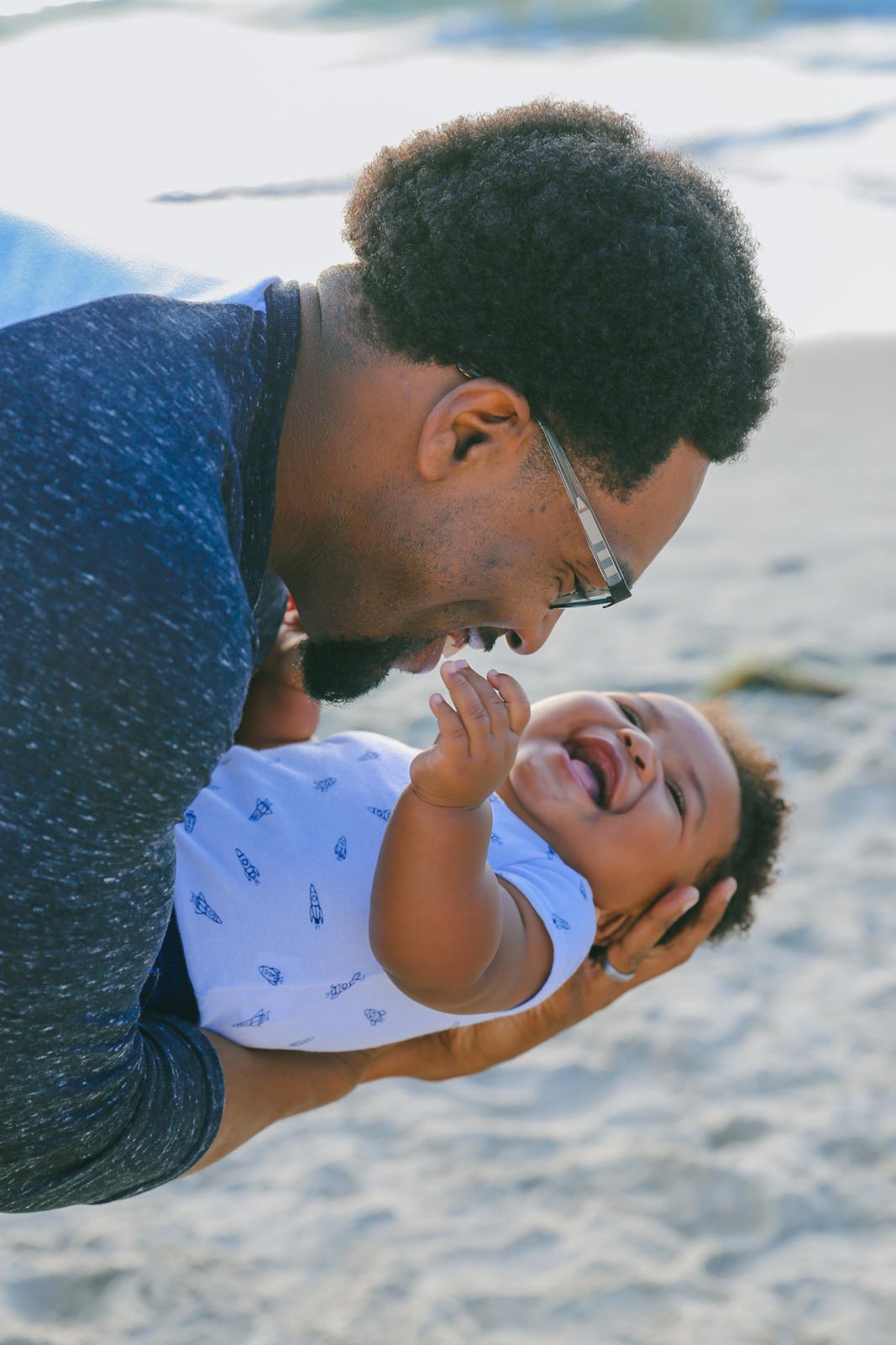 A smiling man in glasses cradling a baby on a beach.