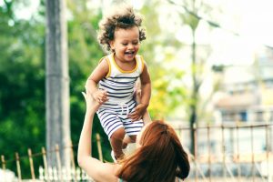 A woman with her back to the camera lifts a smiling young girl above her head.